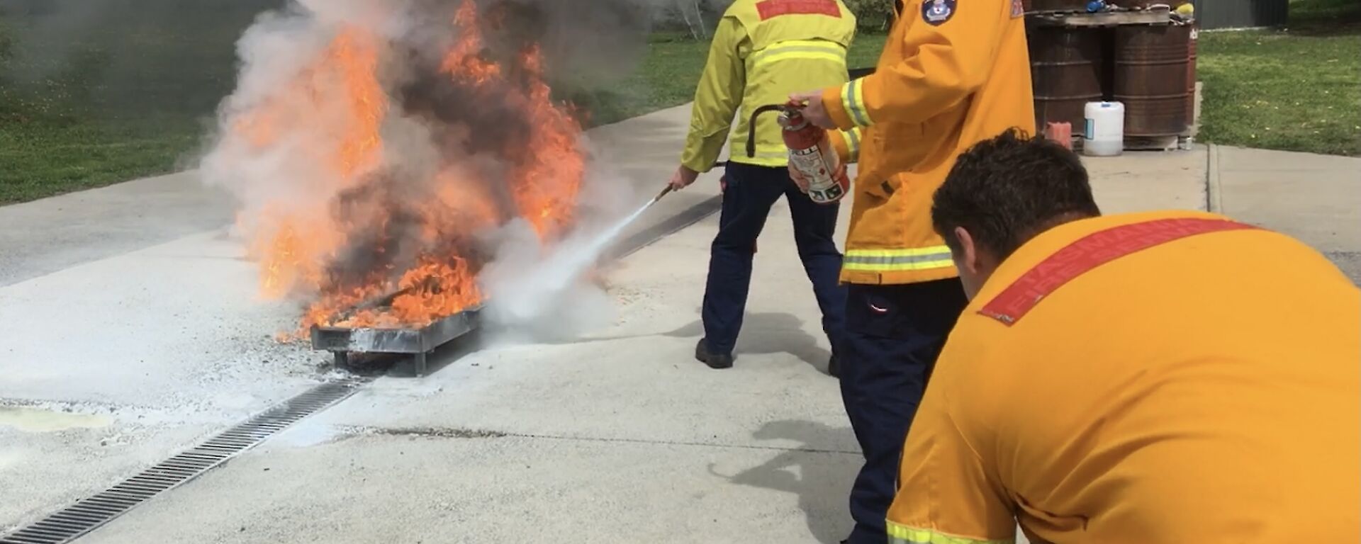 Three men in fire safety uniforms.  On is dousing a large fire with an extinguisher whilst the other two look on.