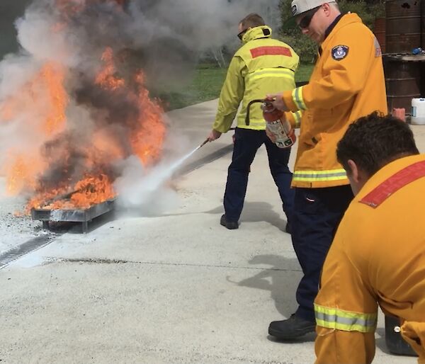 Three men in fire safety uniforms. On is dousing a large fire with an extinguisher whilst the other two look on.