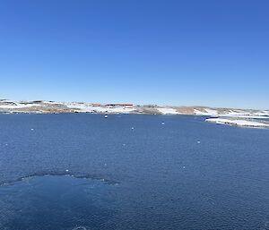 A view across the sea ice from ship towards a snowy shore.  Small buildings can be seen on the shore.