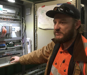 A close up of a bearded man in hi-vis with a baseball cap, standing beside an open circuit box.