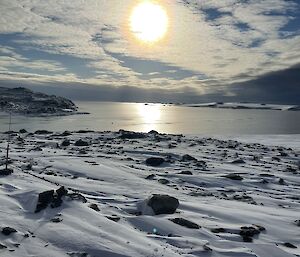 A snow landscape in the foreground, looking out across the sea to the distance.  A large yellow sun in the cloudy sky.