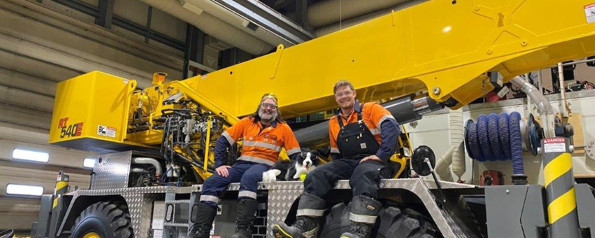 Two expeditioner sitting on the yellow crane with a stuffed toy Border Collie dog in the middle.