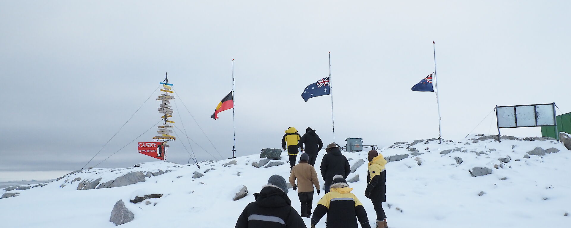 A group of expeditioners walk up a snowy slope towards 3 flag poles flying the Australian, New Zealand and Aboriginal flags at half mast