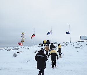 A group of expeditioners walk up a snowy slope towards 3 flag poles flying the Australian, New Zealand and Aboriginal flags at half mast