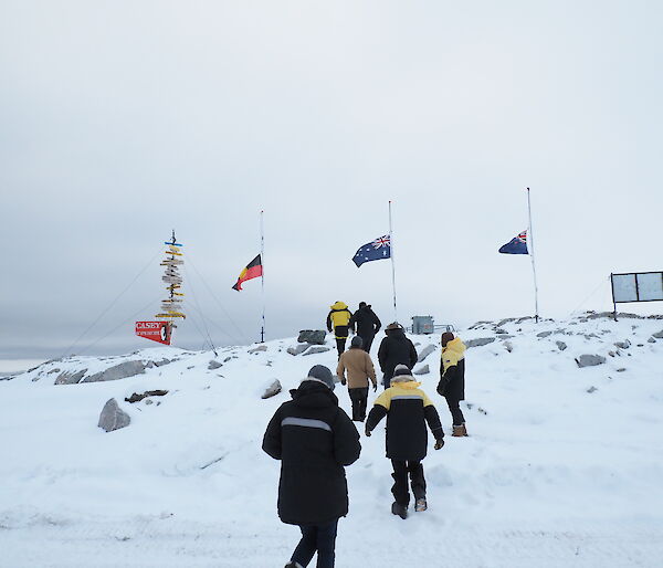 A group of expeditioners walk up a snowy slope towards 3 flag poles flying the Australian, New Zealand and Aboriginal flags at half mast