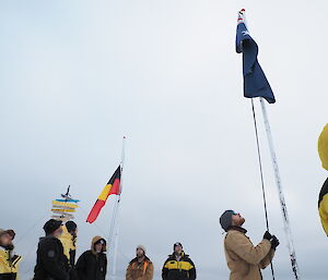 A group of people watch as one expeditioner raises the Australian flag up one of two flagpoles.  The other pole has the Aboriginal flag at half mast.