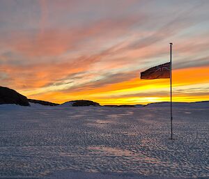 Bright orange sky reflecting on the sea ice with the Australian flag at half-mast in the foreground.