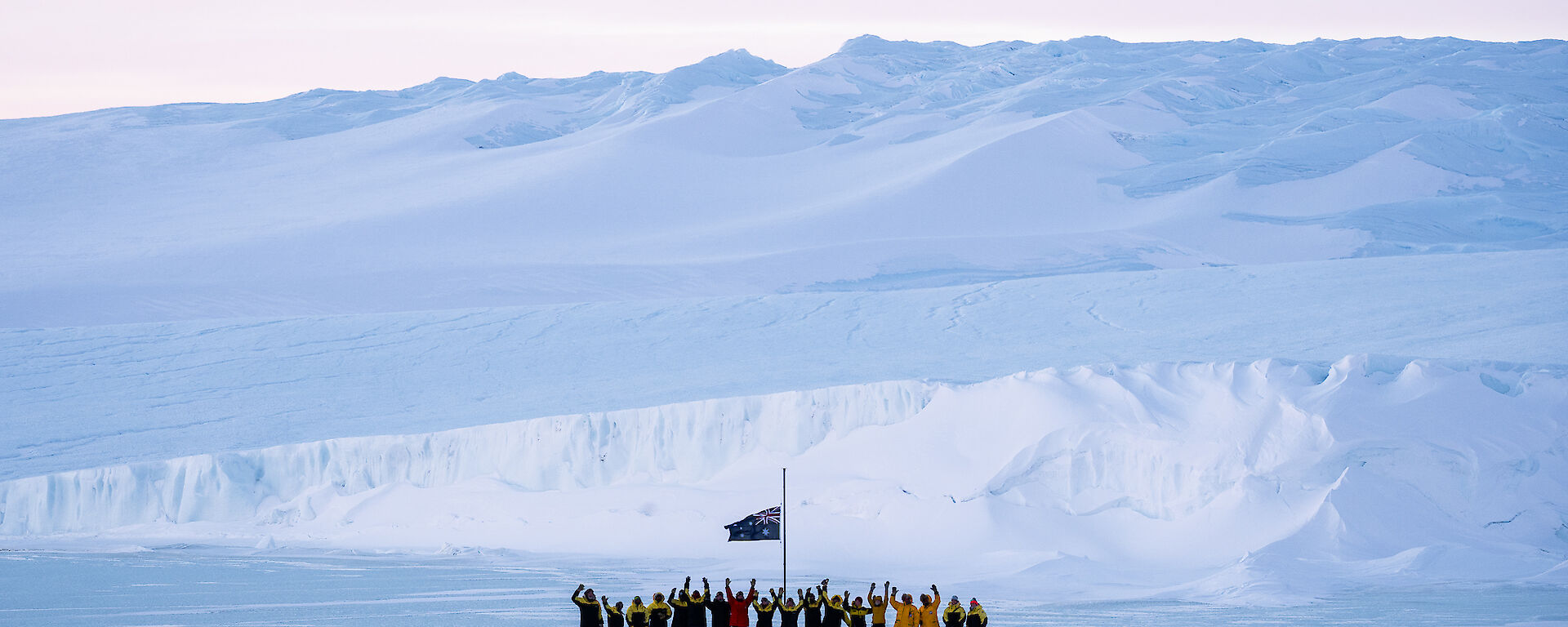 Mawson expeditioners with arms in the air standing at the flag pole with the plateau rising up from the sea ice in the background.