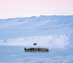 Mawson expeditioners with arms in the air standing at the flag pole with the plateau rising up from the sea ice in the background.