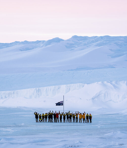 Mawson expeditioners with arms in the air standing at the flag pole with the plateau rising up from the sea ice in the background.