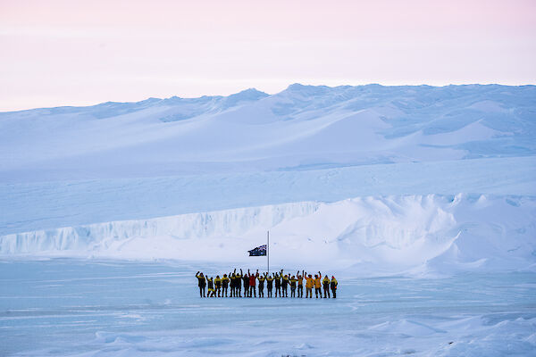 Mawson expeditioners with arms in the air standing at the flag pole with the plateau rising up from the sea ice in the background.