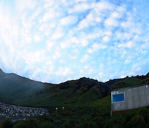 A field hut in the green tussocks of the hillside with a colony of penguins on the beach below