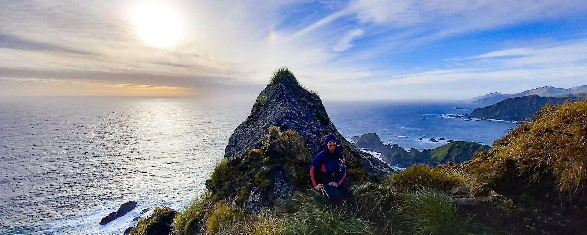 An expeditioner resting in the grassy tussocks against a large pointed rock with the sea and blue sky behind it