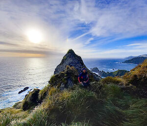 An expeditioner resting in the grassy tussocks against a large pointed rock with the sea and blue sky behind it