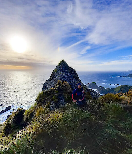 An expeditioner resting in the grassy tussocks against a large pointed rock with the sea and blue sky behind it