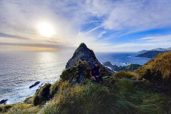 An expeditioner resting in the grassy tussocks against a large pointed rock with the sea and blue sky behind it