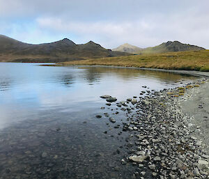 The pebbly shore of a lake with green hills in the background