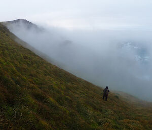 An expeditioner heading down a steep green slope towards the mist below