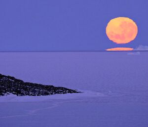 A large pink supermoon in the sky above a snowy landscape with an iceberg on the horizon