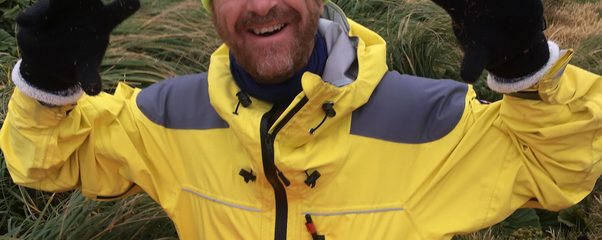 A man in bright yellow cold weather clothing, gloves and a bright hat holds his hands up to camera and smiles