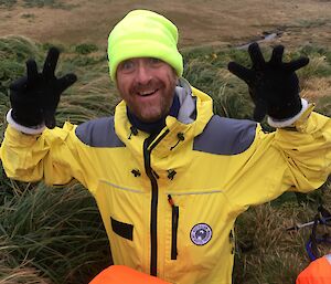 A man in bright yellow cold weather clothing, gloves and a bright hat holds his hands up to camera and smiles