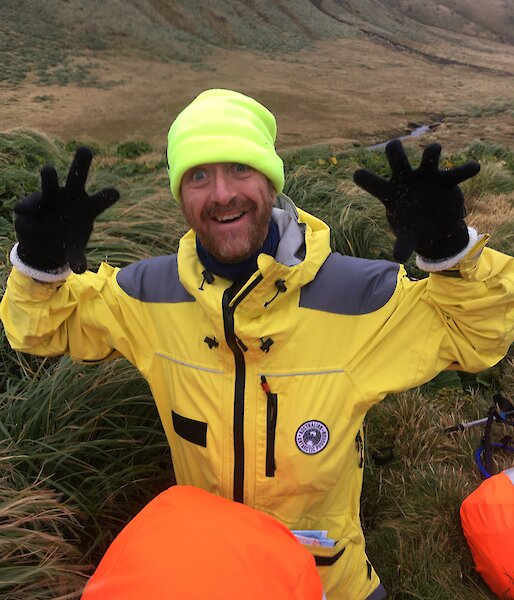A man in bright yellow cold weather clothing, gloves and a bright hat holds his hands up to camera and smiles