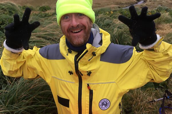 A man in bright yellow cold weather clothing, gloves and a bright hat holds his hands up to camera and smiles