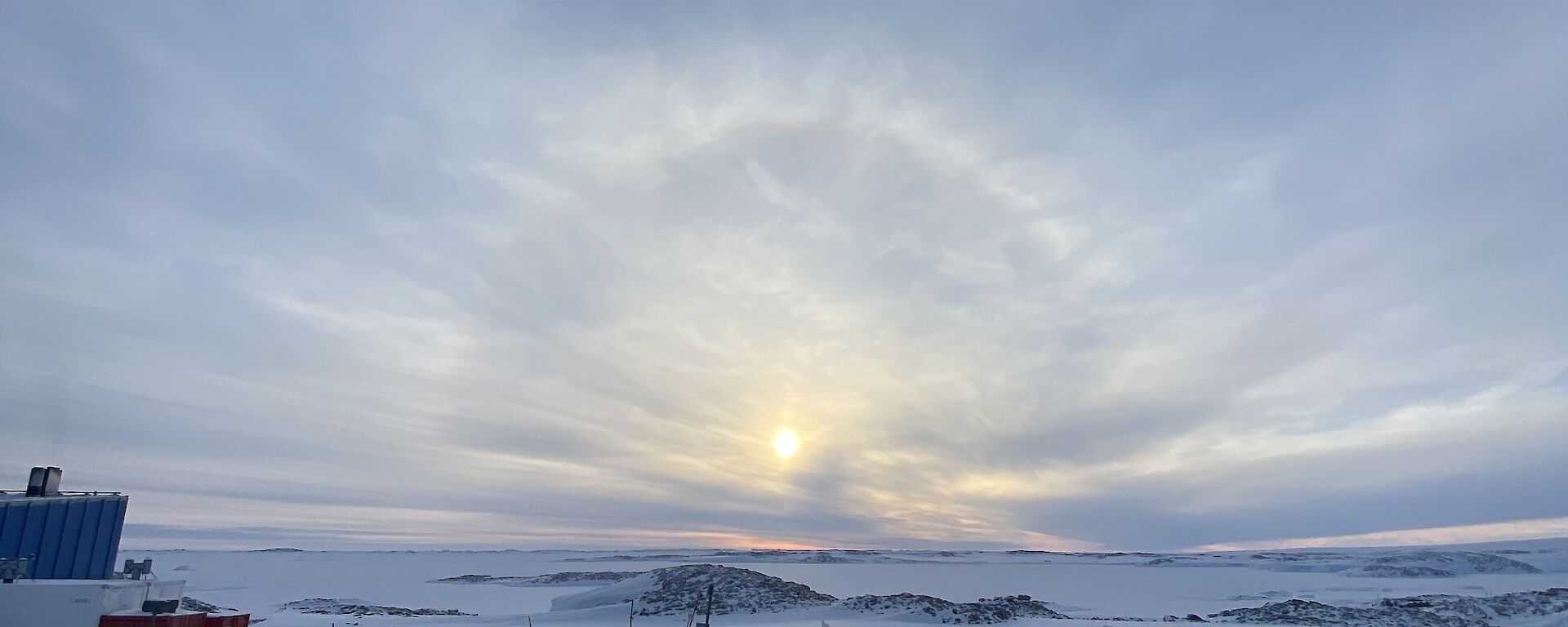 A snowy scene with a cloudy sky in the distance.  A sun halo is peeping through the cloud.
