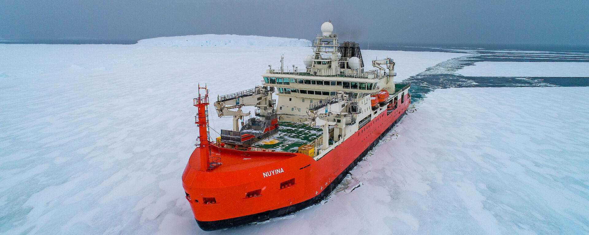 An aerial drone photo of an orange and white ship breaking through sea ice. There is a long channel of water behind the ship where it has broken a path through the white expanse of the ice.