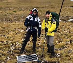 Two men stand in a grass covered landscape with a seismometer on the ground in front of them.  One carries a large backpack with an antaena sticking out of it.