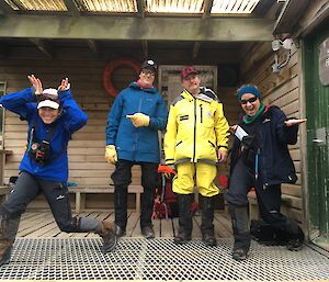 Four people stand in front of a shed like building,  two are striking goofy poses.