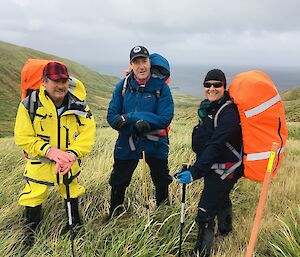 Three expeditioners stand at the top of a hill in bright coloured walking gear and backpacks