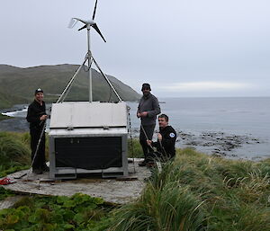Three expeditioners standing next to a small hut and a small wind turbine
