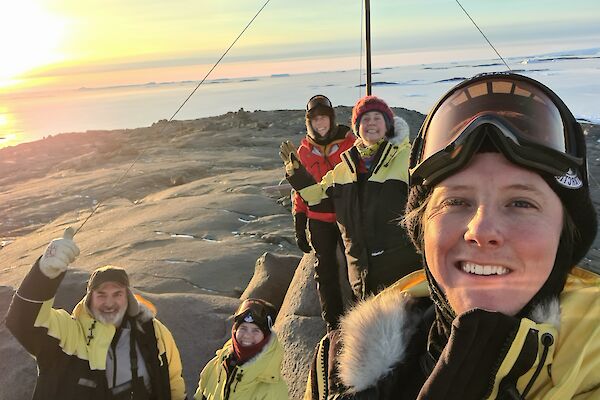 5 Expeditioners on top of the highest point on Beche Island posing for camera with rising sun in the background
