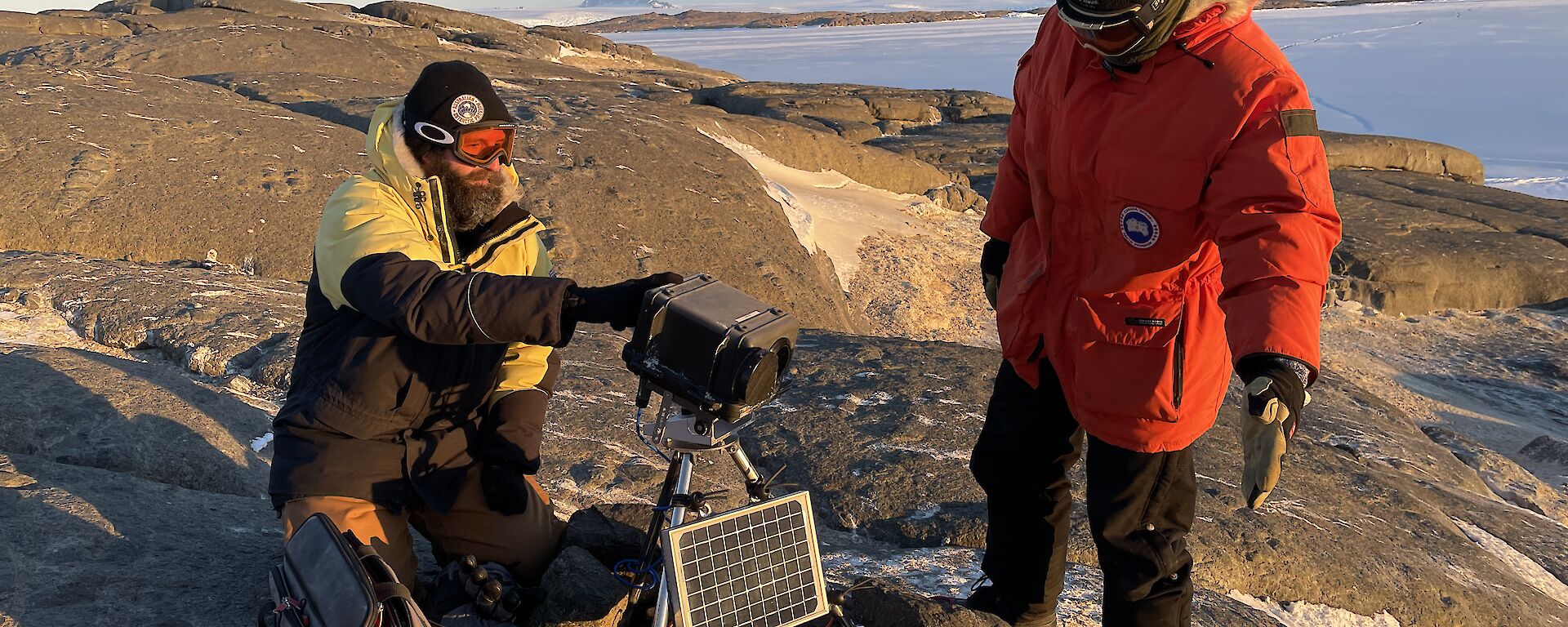 Two expeditioners working on a remote bird camera on top of a rocky hill