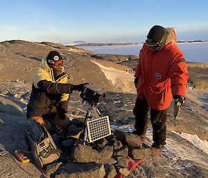 Two expeditioners working on a remote bird camera on top of a rocky hill