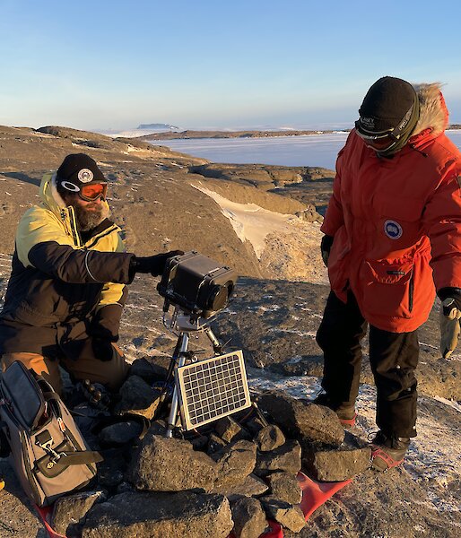 Two expeditioners working on a remote bird camera on top of a rocky hill