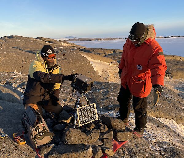 Two expeditioners working on a remote bird camera on top of a rocky hill