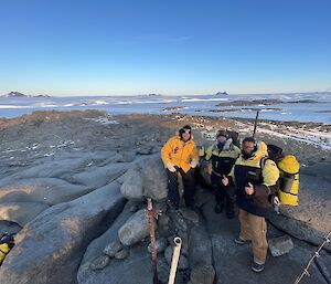 Three expeditioners standing on a rocky hill top with sea ice, the ice plateau and mountain in the background.