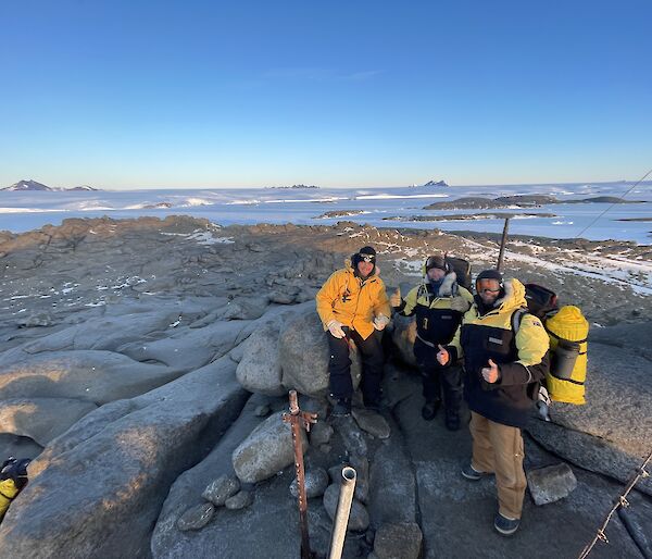 Three expeditioners standing on a rocky hill top with sea ice, the ice plateau and mountain in the background.