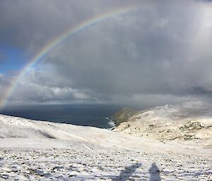 Looking across some snow covered ground to the sea with a rainbow overhead