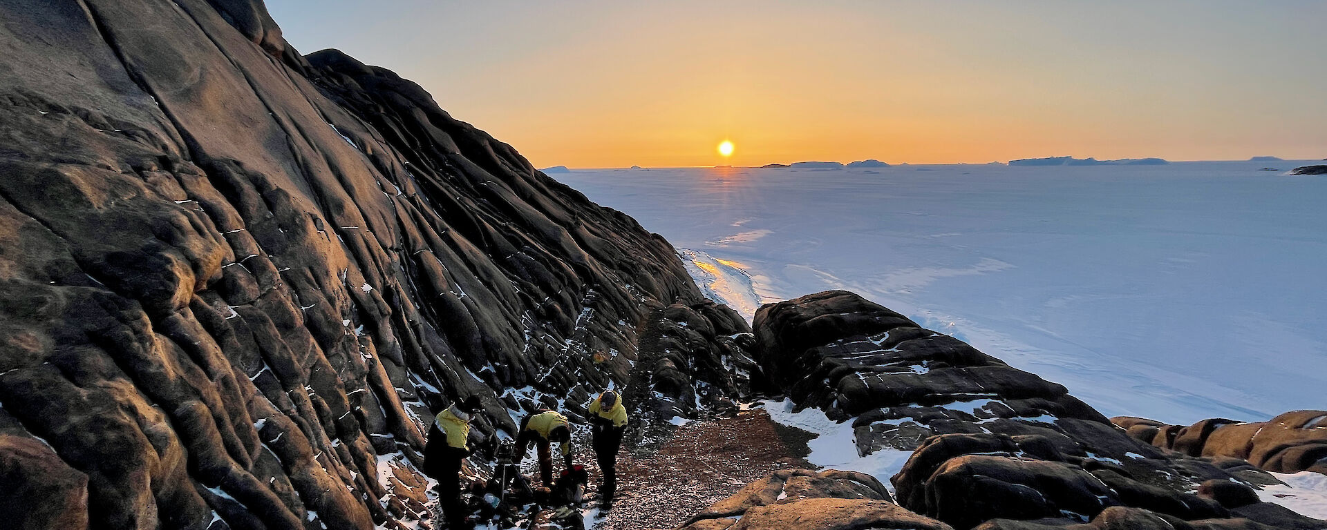 Three expeditioners working on a rocky ledge with the sun producing an orange glow as it goes down in the background.