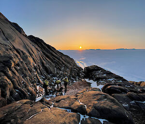 Three expeditioners working on a rocky ledge with the sun producing an orange glow as it goes down in the background.