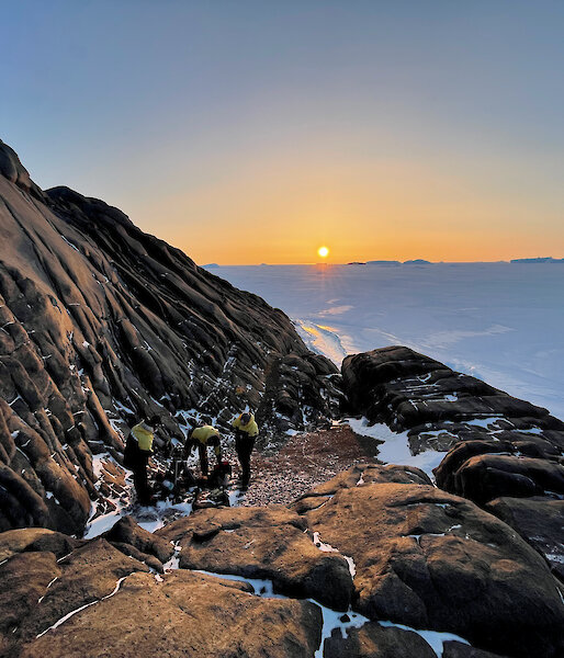 Three expeditioners working on a rocky ledge with the sun producing an orange glow as it goes down in the background.