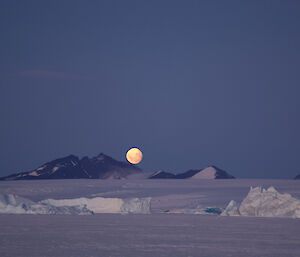 The moonrise over a mountain range with the ice plateau and sea ice in the foreground.