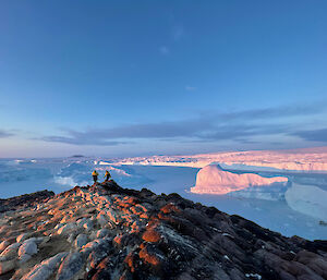 Two expeditioners on top a rocky island with icebergs locked in the sea ice in the background.