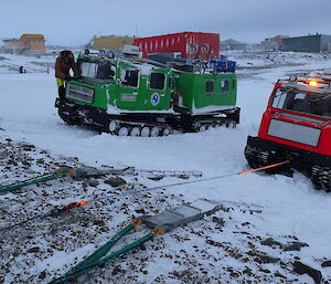 Two Hagglands sitting in the snow.  One is attached by rope to a winch with a ramp in front of it.