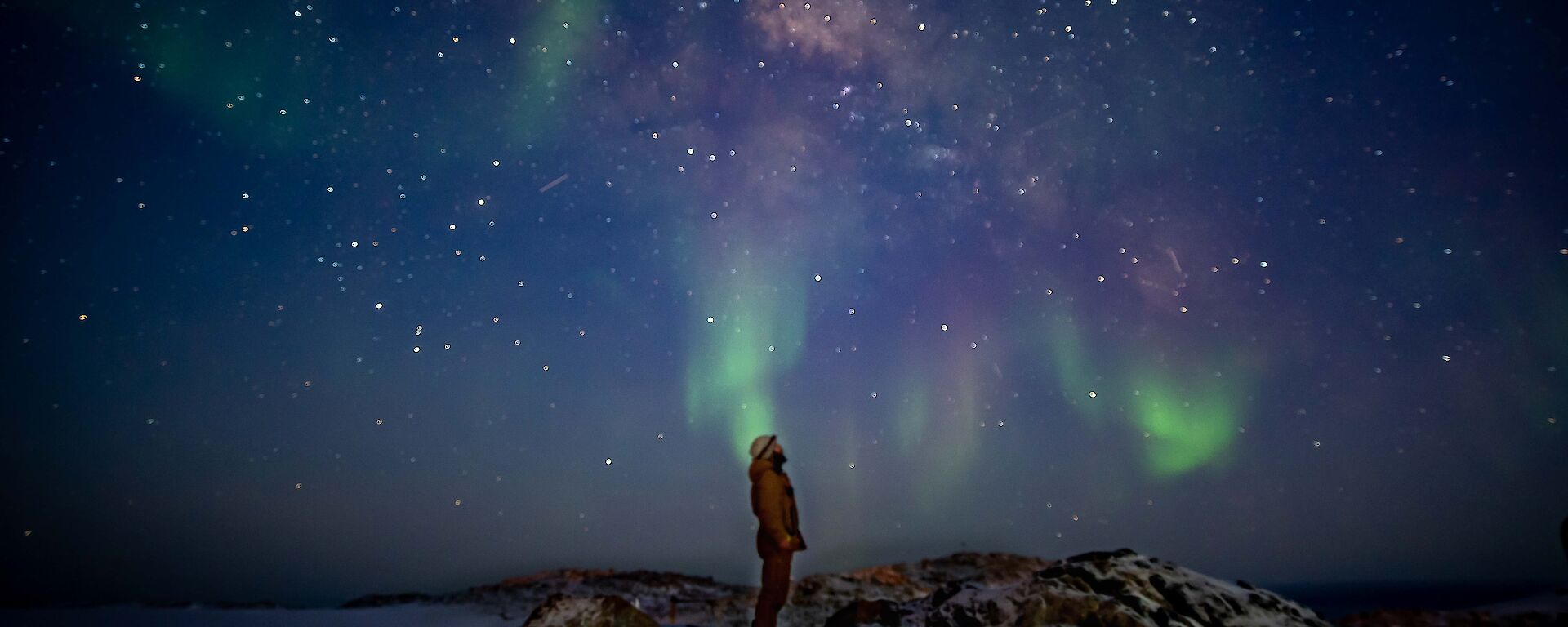 A man stands silhouetted against the sky.  A green and pink aurora and stars light up the sky.