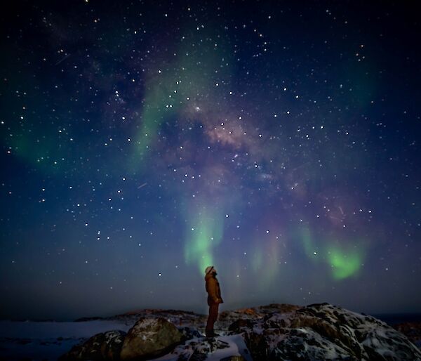 A man stands silhouetted against the sky.  A green and pink aurora and stars light up the sky.