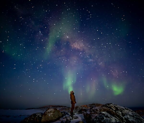 A man stands silhouetted against the sky. A green and pink aurora and stars light up the sky.